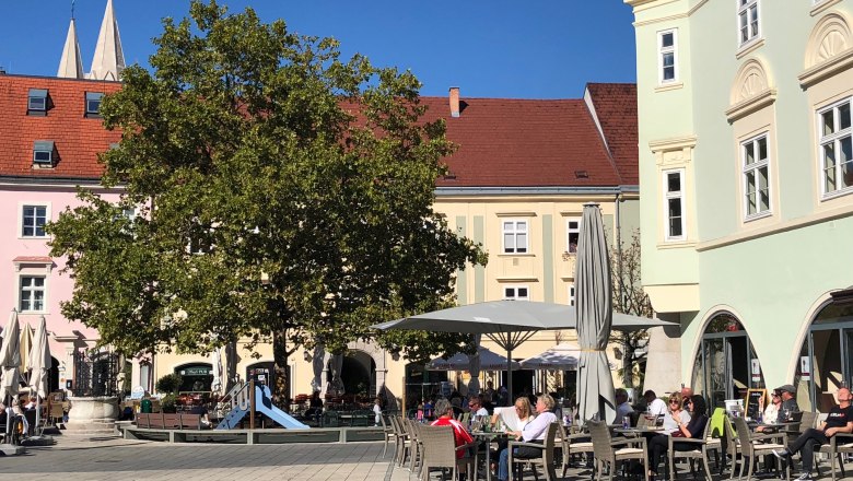 Sunny main square with a tree and playground at Eterno. People sit outside and enjoy the weather.