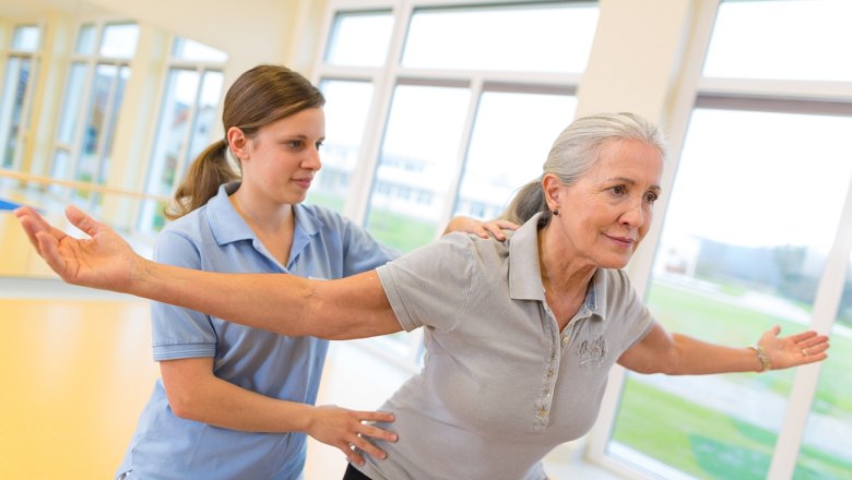 An older woman does gymnastic exercises with the support of a younger woman in a bright room.