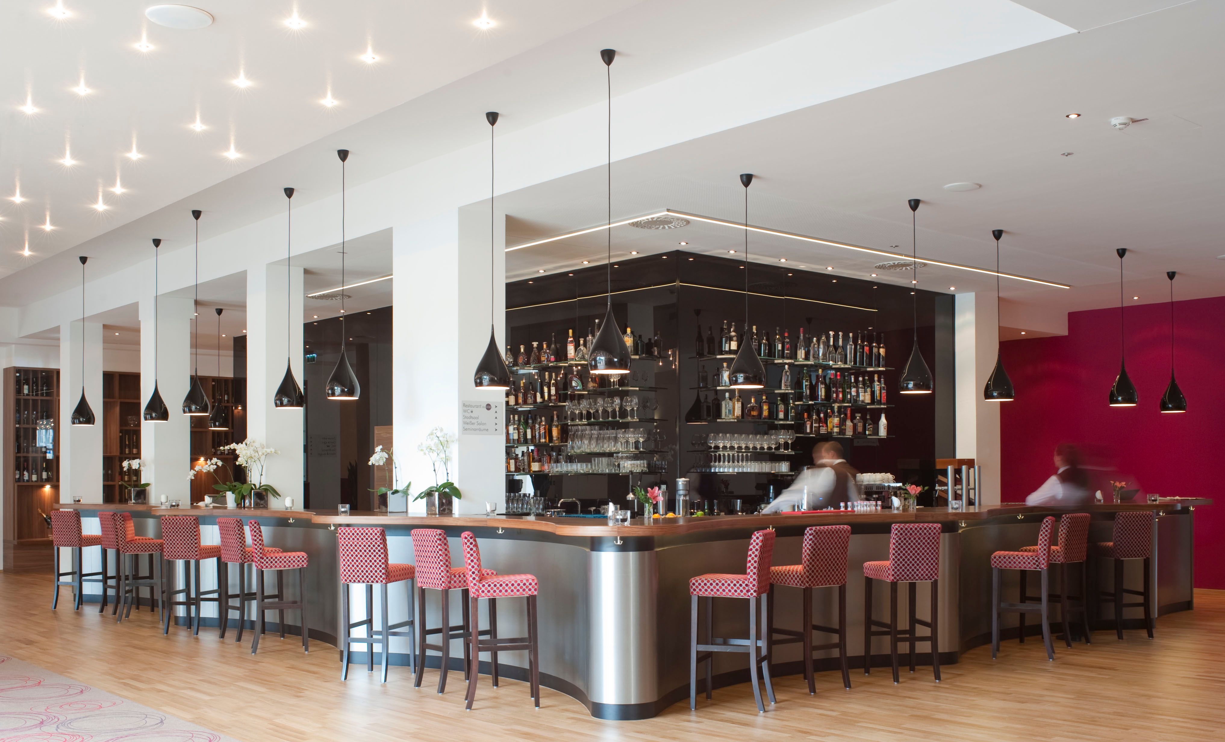 Modern hotel bar with red chairs and black hanging lamps.