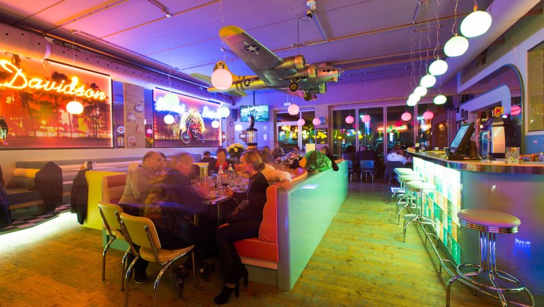Interior view of a colorfully lit diner with bar, tables and an airplane model on the ceiling.