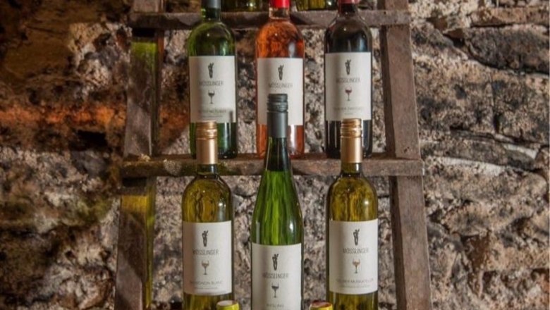 Wine bottles on a wooden shelf in front of a stone wall.