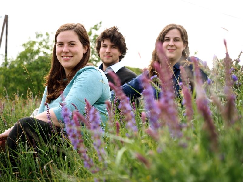 Three young adults sit in a flowering field.