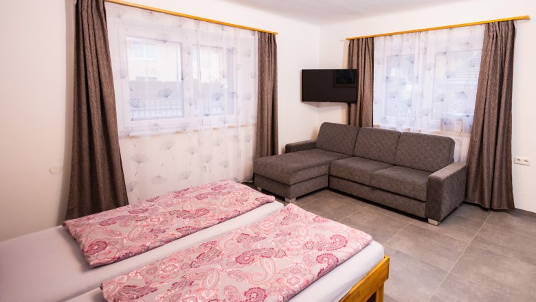 A bedroom with a double bed, gray sofa, two windows with curtains and a TV on the wall.