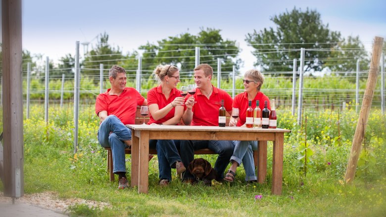 Four people in red shirts sit at an outdoor table with bottles of wine and glasses, surrounded by vines.