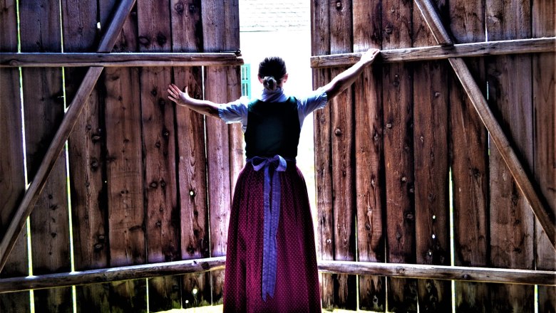 Höblinghof, © Gluschitz-Goebel A woman in traditional dress opens large wooden doors to the outside.