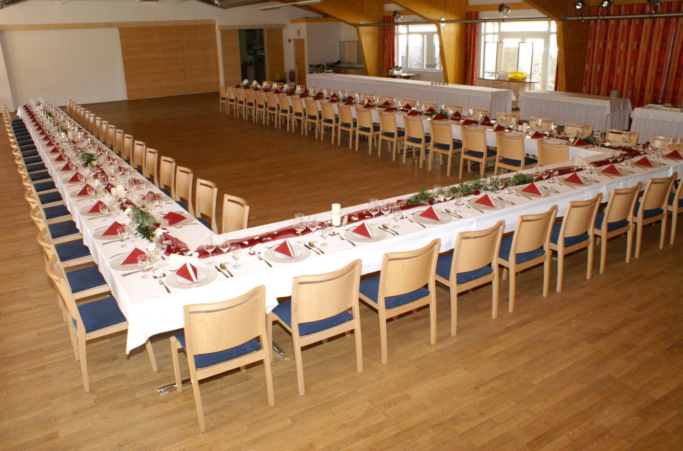 A large ballroom with tables set in a U-shape, decorated with red napkins and flowers.