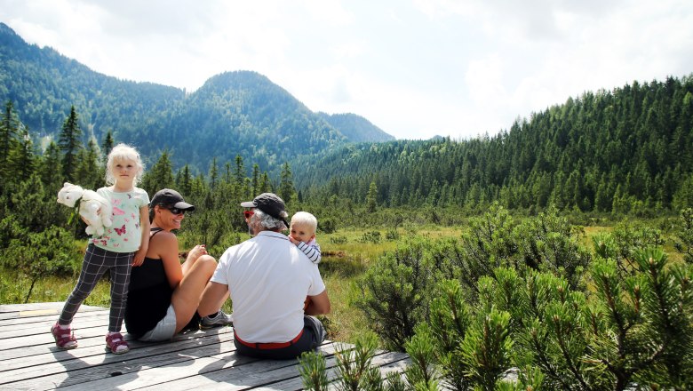 Family sitting on a wooden platform in the Leckermoos high moor, surrounded by mountains and forest.