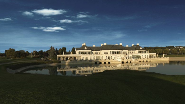 An elegant clubhouse is reflected in a tranquil lake, surrounded by green lawns and trees under a clear blue sky.
