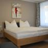 Wooden double bed with white bed linen. Behind it a window with curtains and a painting.