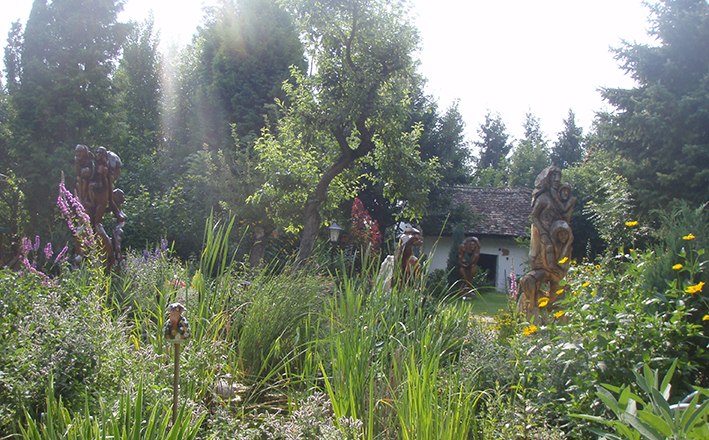 A lush garden with sculptures, trees and a small building in the background.