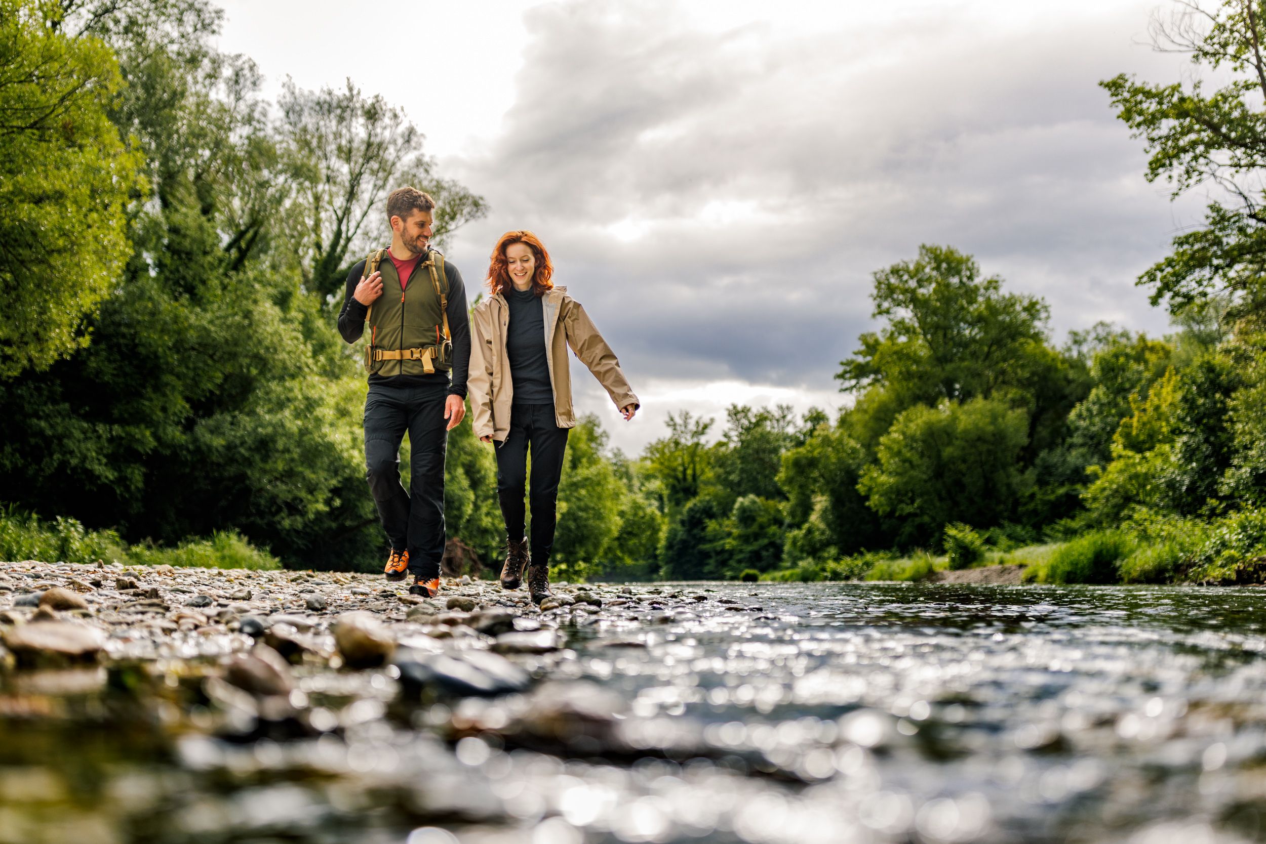 A woman and a man stroll happily along the gravel bed next to the Leitha, green forest in the background