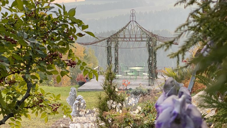 A romantic garden with gazebo, green chairs and table, surrounded by trees and flowers.