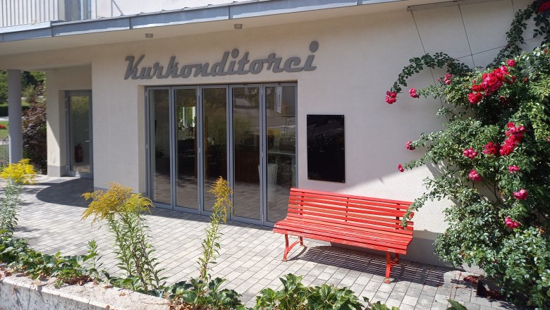 Exterior view of a building with the inscription 'Kurkonditorei', red bench and blooming roses.