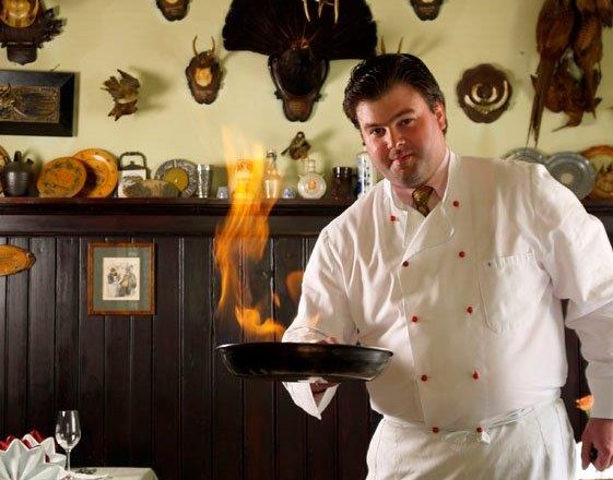 A chef in a white uniform flamb&eacute;es a dish in a rustic kitchen with hunting trophies on the wall.