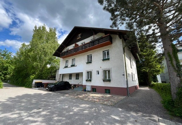 Two-storey house with white façade and dark roof, surrounded by trees and a paved courtyard.