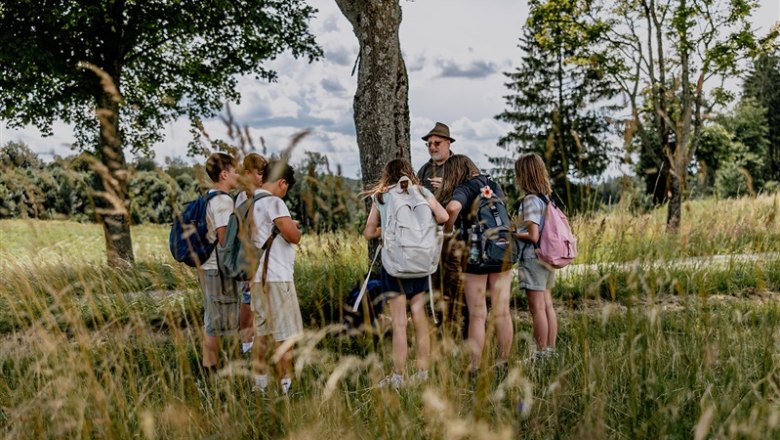 Group of children with backpacks and an adult outdoors, surrounded by trees and grass.