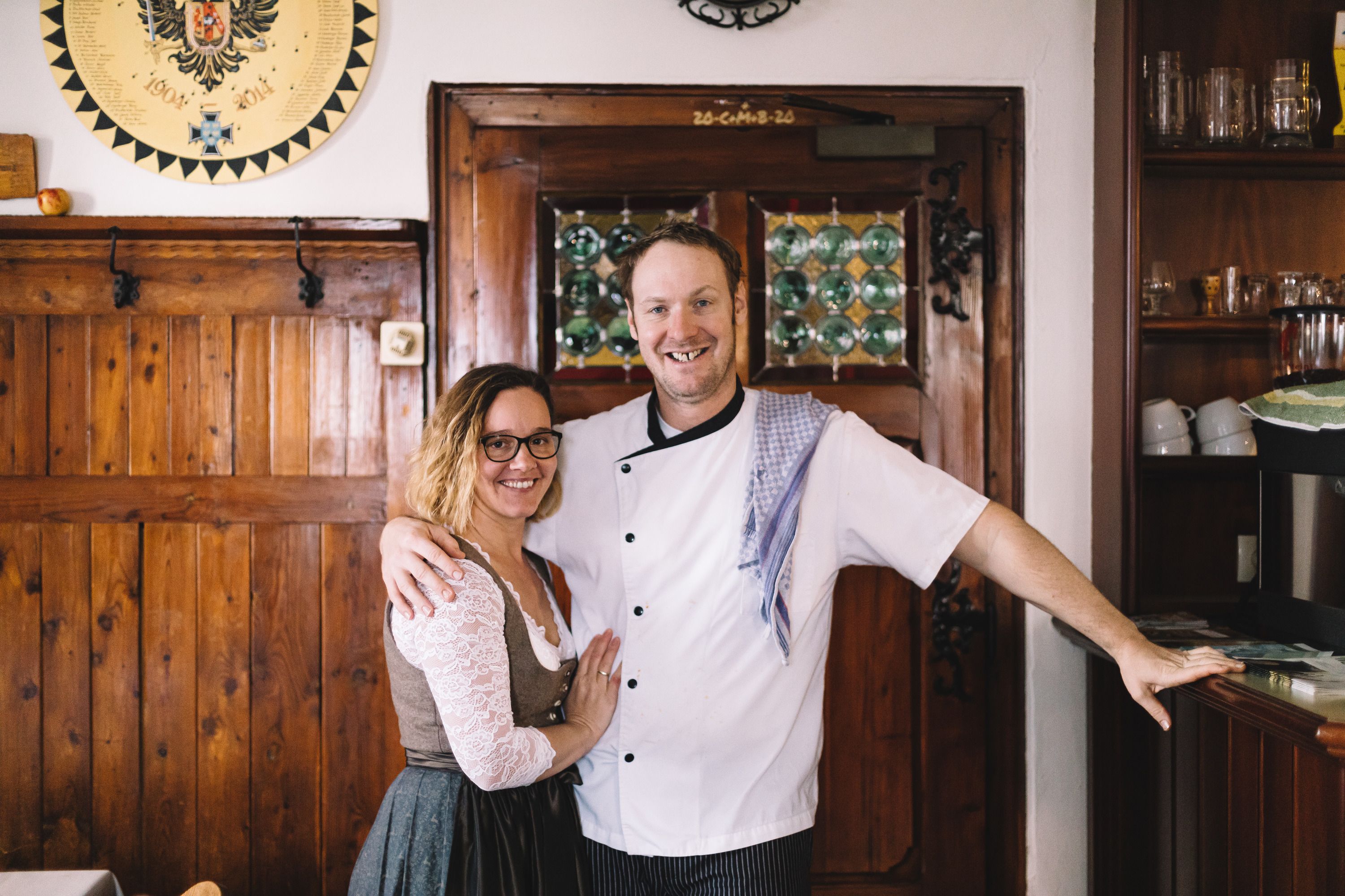 A man in chef's clothes and a woman in traditional dress stand smiling in a rustic room.