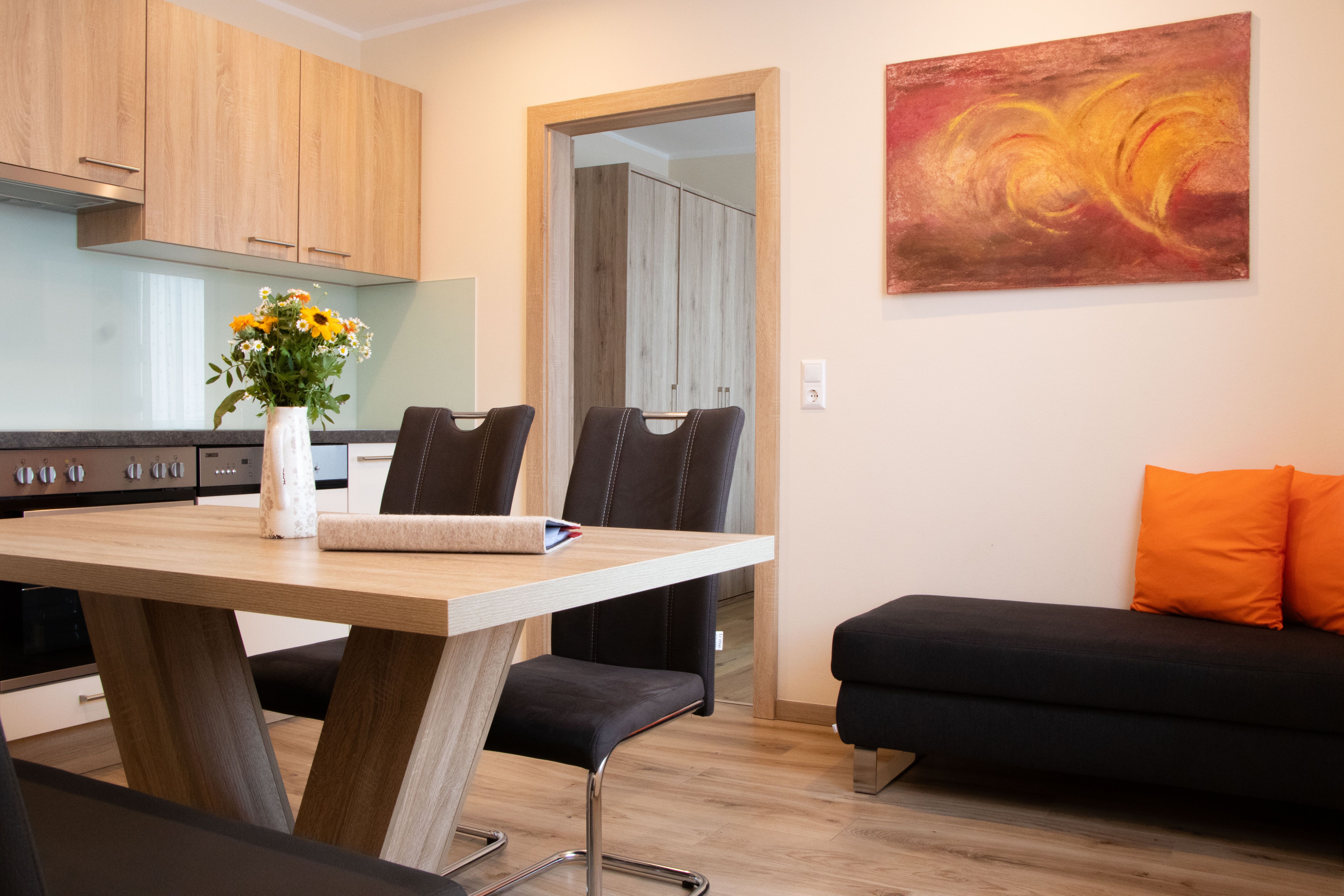 Modern kitchen with wooden furniture, a dining table, chairs, and a sofa with orange cushions.