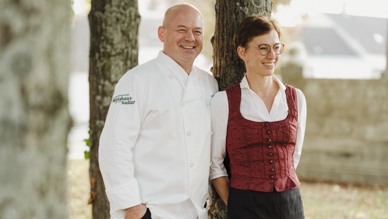 A man in a white chef's jacket and a woman in traditional dress stand smiling by a tree.