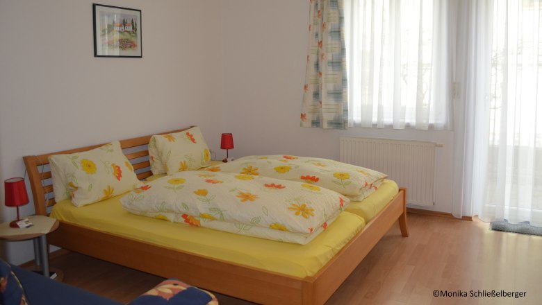 Double room, © Monika Schließelberger A cozy double room with a wooden bed, colorful bedding and two red bedside lamps. A window with curtains is in the background.