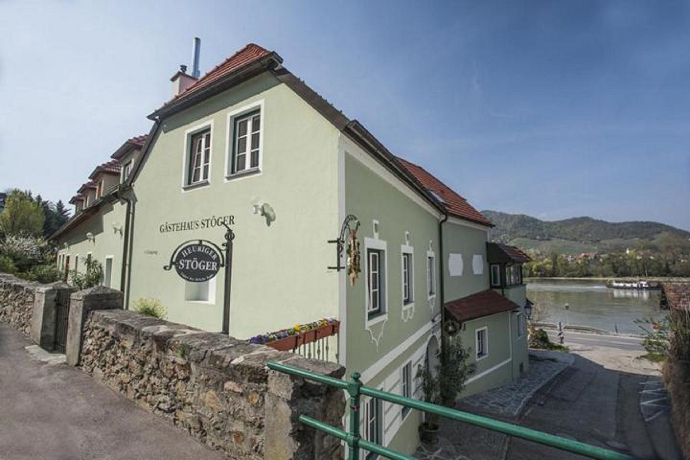 A green guest house with red roofs on the riverbank, surrounded by hills and flowering plants.