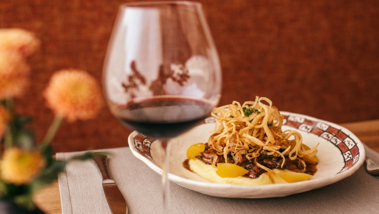 A plate of glazed veal liver on mashed potatoes, garnished with fried onions, next to a glass of red wine on a table.