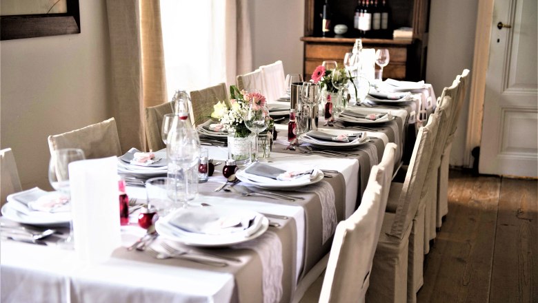Elegantly laid table with white tablecloths, flower arrangements and crockery in a room with a wooden floor.