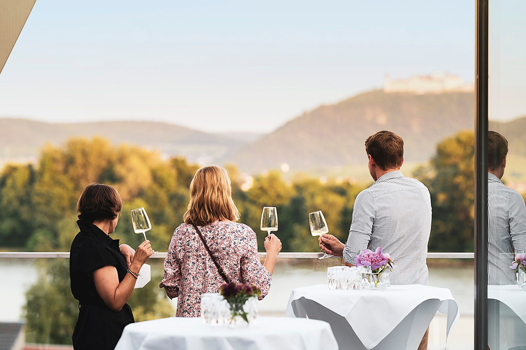 Three people on a terrace with wine glasses, view of the hills and river.