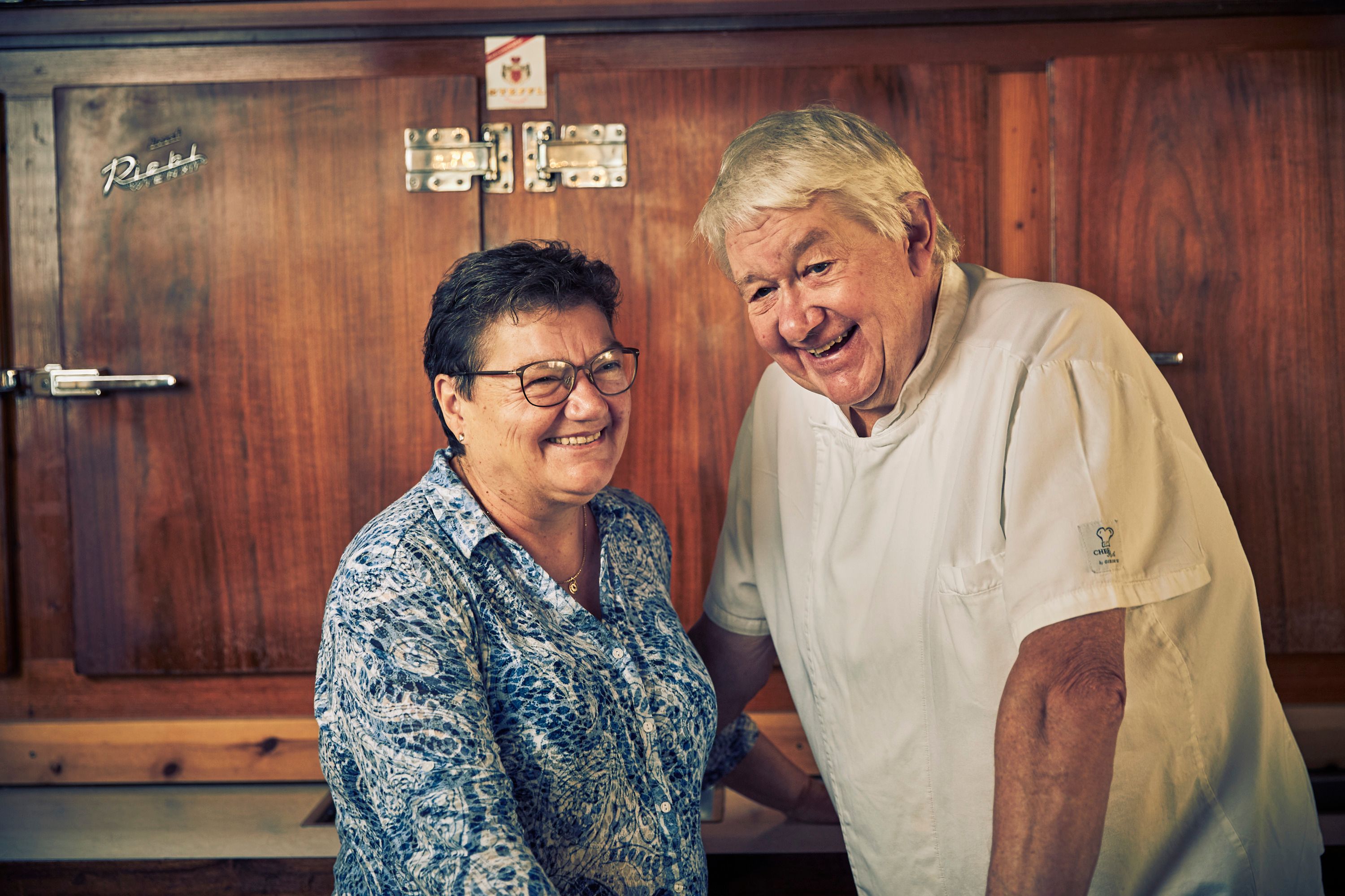 An elderly couple smiles in a kitchen. The man is wearing a chef's jacket, the woman a patterned blouse. They are standing in front of a wooden cupboard.