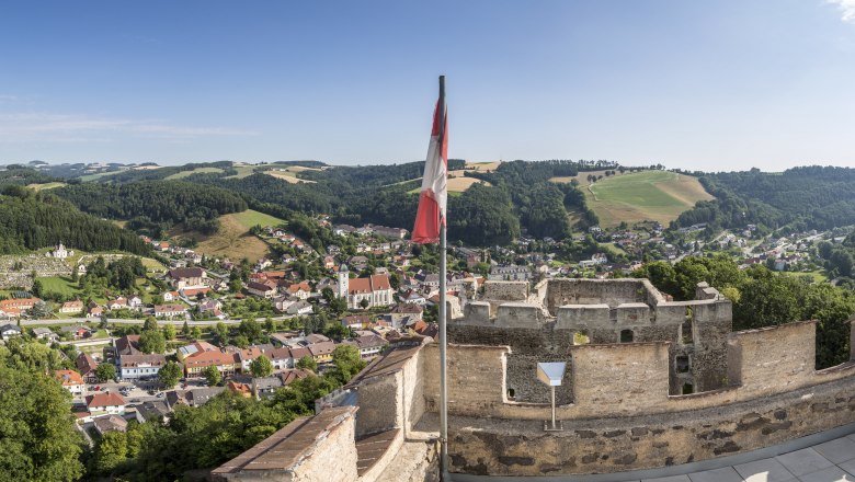 Panoramic view from a ruined castle to a town with hills in the background and an Austrian flag in the foreground.
