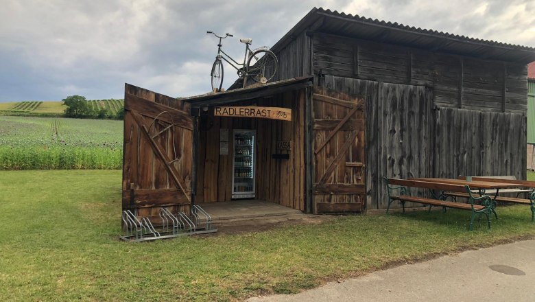 A rustic wooden hut with the sign 'Radlerrast' and a bicycle on the roof, surrounded by fields and a bench.