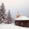A small church in the snow with fir trees in the background.