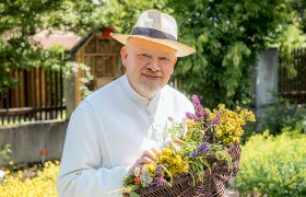 A man in a white robe and straw hat holds a basket of colorful herbs in a garden.