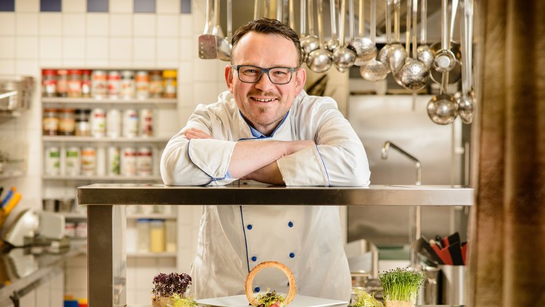 A chef in a white uniform leans on a stainless steel counter in a kitchen. In front of him is a plate of decorative food.