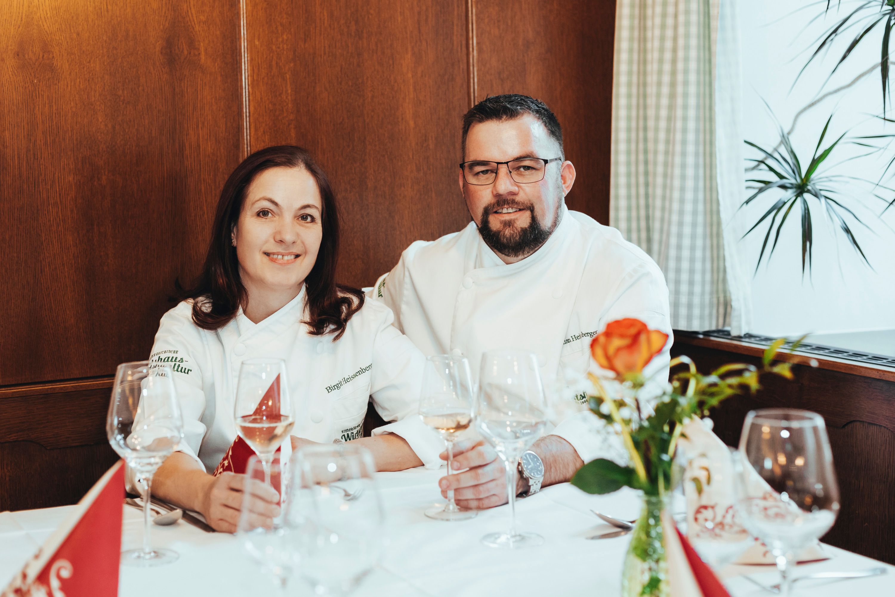 Two people in chef's jackets sit at a laid table with wine glasses.