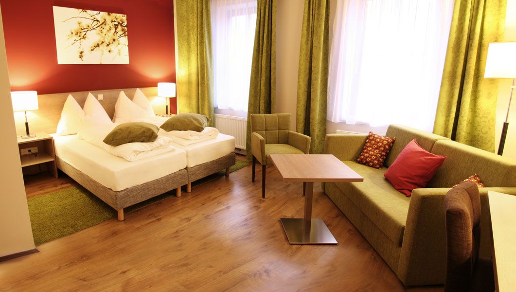 Cozy hotel room with double bed, green couch, table and chair. Warm colors and modern furnishings.