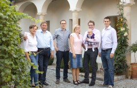 A family stands outside in front of a building with arches, surrounded by plants.