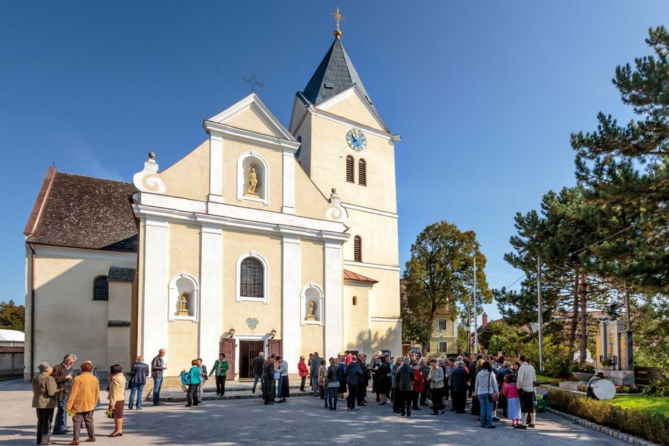 People gather in front of a church with a tower and clock in sunny weather.