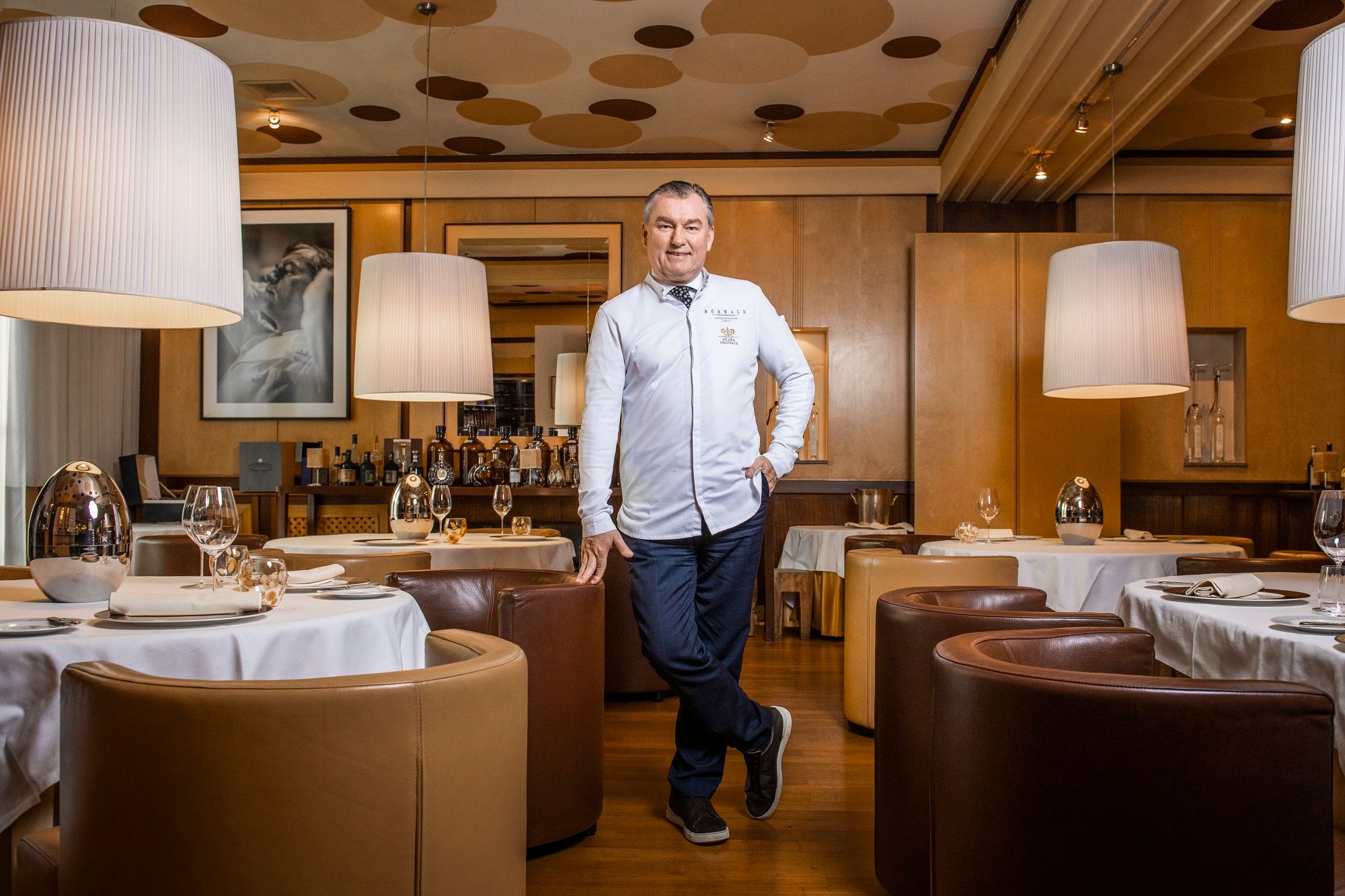 A man in a white chef's jacket stands in an elegant restaurant with round tables and large lamps.
