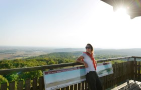 Woman with sunglasses leaning against a railing on a lookout tower with a view of a vast landscape.