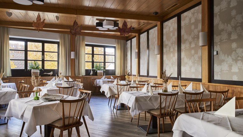An elegant restaurant with wooden furniture, white tablecloths and large windows showing autumnal trees.