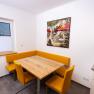Dining area with wooden table, yellow corner bench and chairs, picture on the wall.