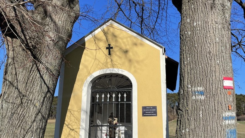 Small yellow chapel between two large trees with a blue sky in the background.