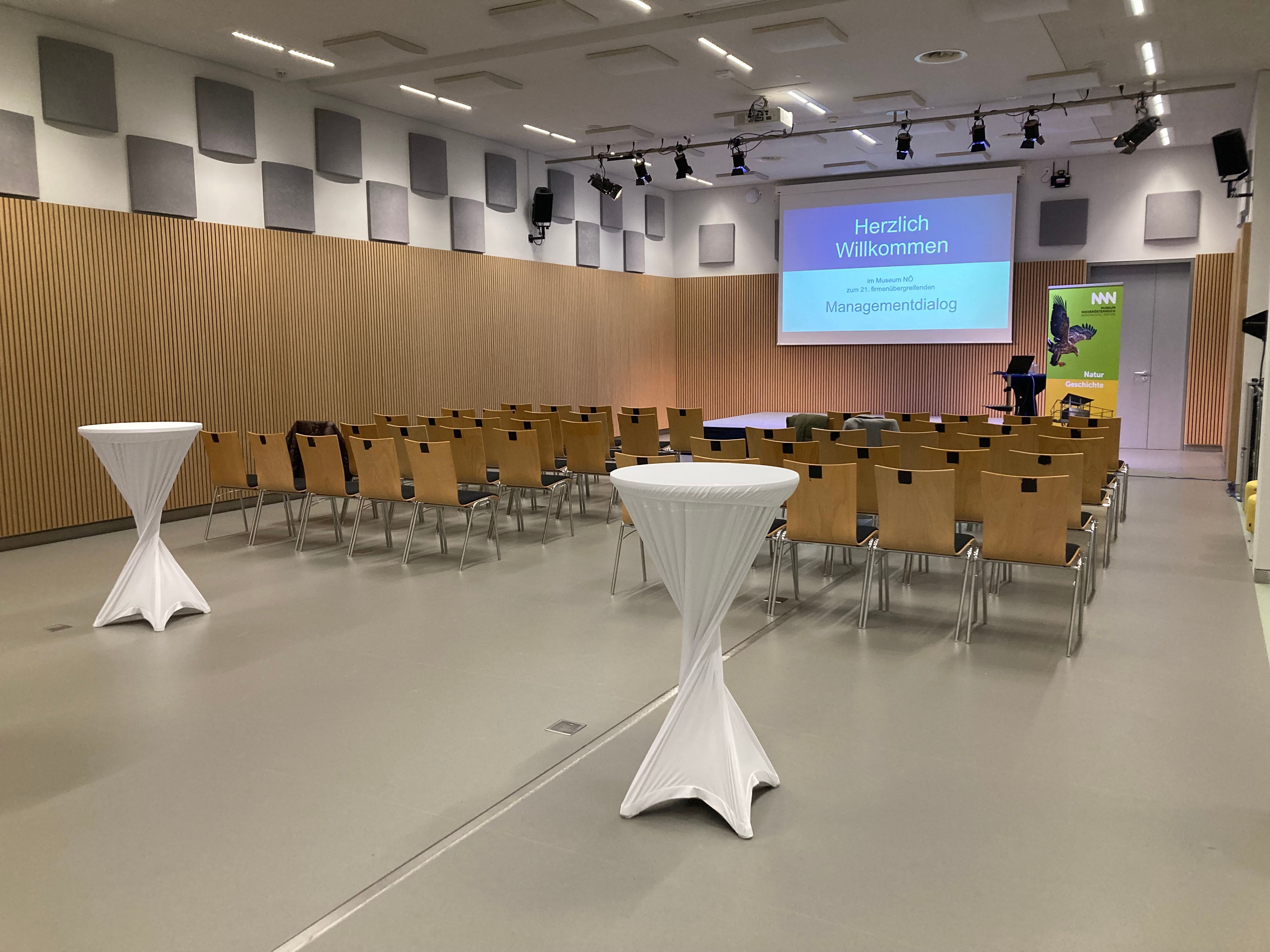 Conference room with rows of chairs, bar tables and a screen with the inscription 'Welcome'.