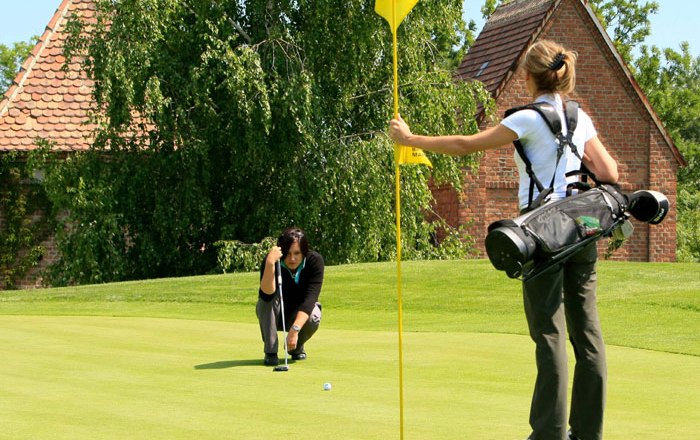 Two women playing golf on a green golf course, one holding the flag, the other aiming at the hole.