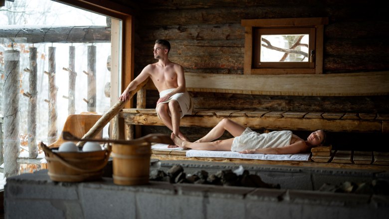 Two people in a wooden sauna, one sitting, the other lying down, both wrapped in towels.