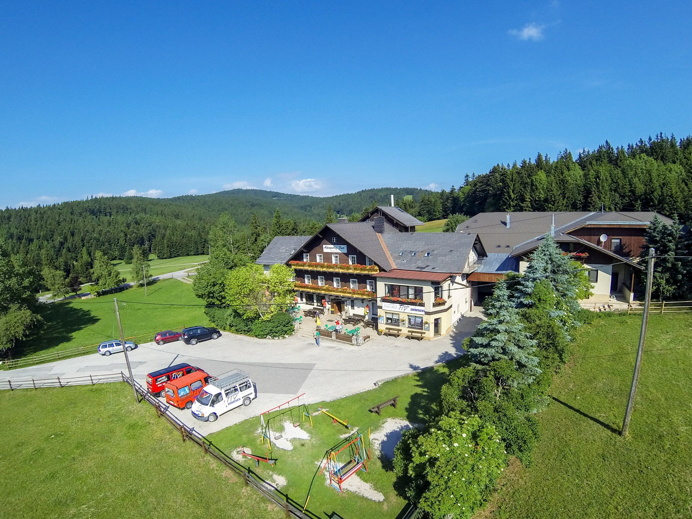 Alpengasthof Postl in a green, wooded landscape with several parked cars and a playground in the foreground.