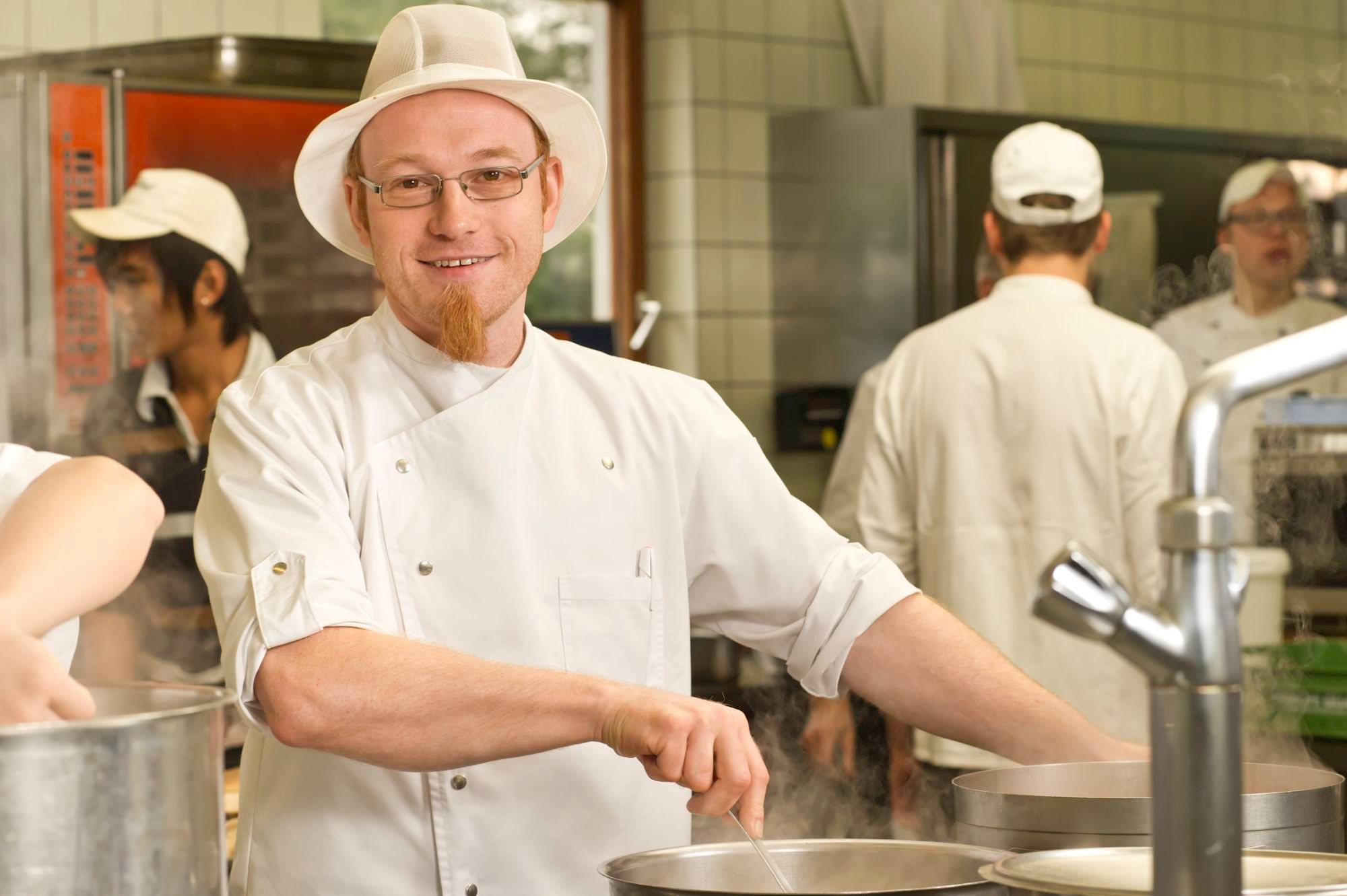 A cook in a white uniform and hat stirs in a pot in a commercial kitchen.