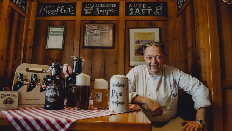 A landlord in traditional dress sits smiling at a table with beer mugs and bottles in a rustic dining room.