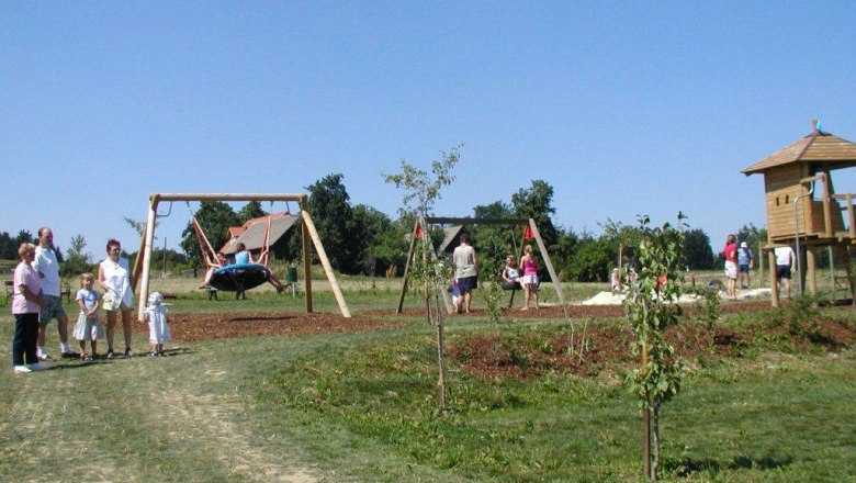 A playground with swings, a climbing frame and children playing. Adults stand next to them. It's a sunny day.